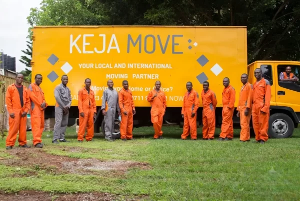 A line of movers in orange and grey uniforms standing outdoors in front of a large yellow moving truck on green grass.