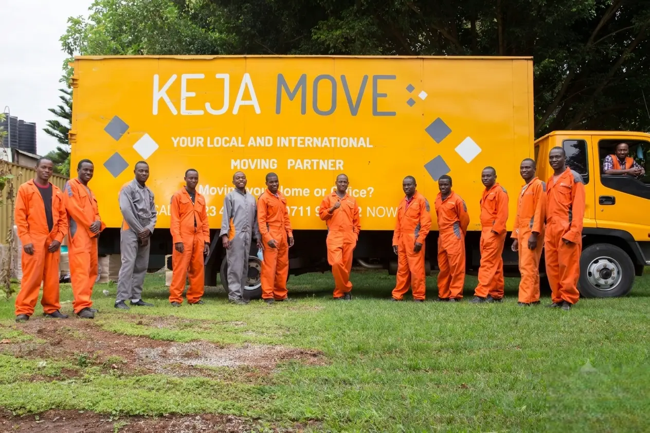 A line of movers in orange and grey uniforms standing outdoors in front of a large yellow moving truck on green grass.